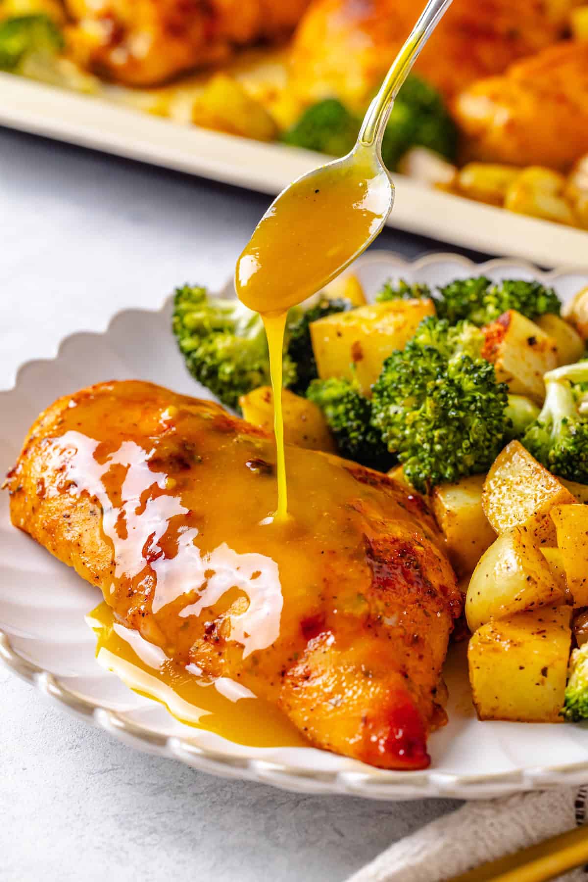 A spoon drizzling honey mustard sauce over a roasted chicken breast on a white plate alongside potatoes and broccoli.