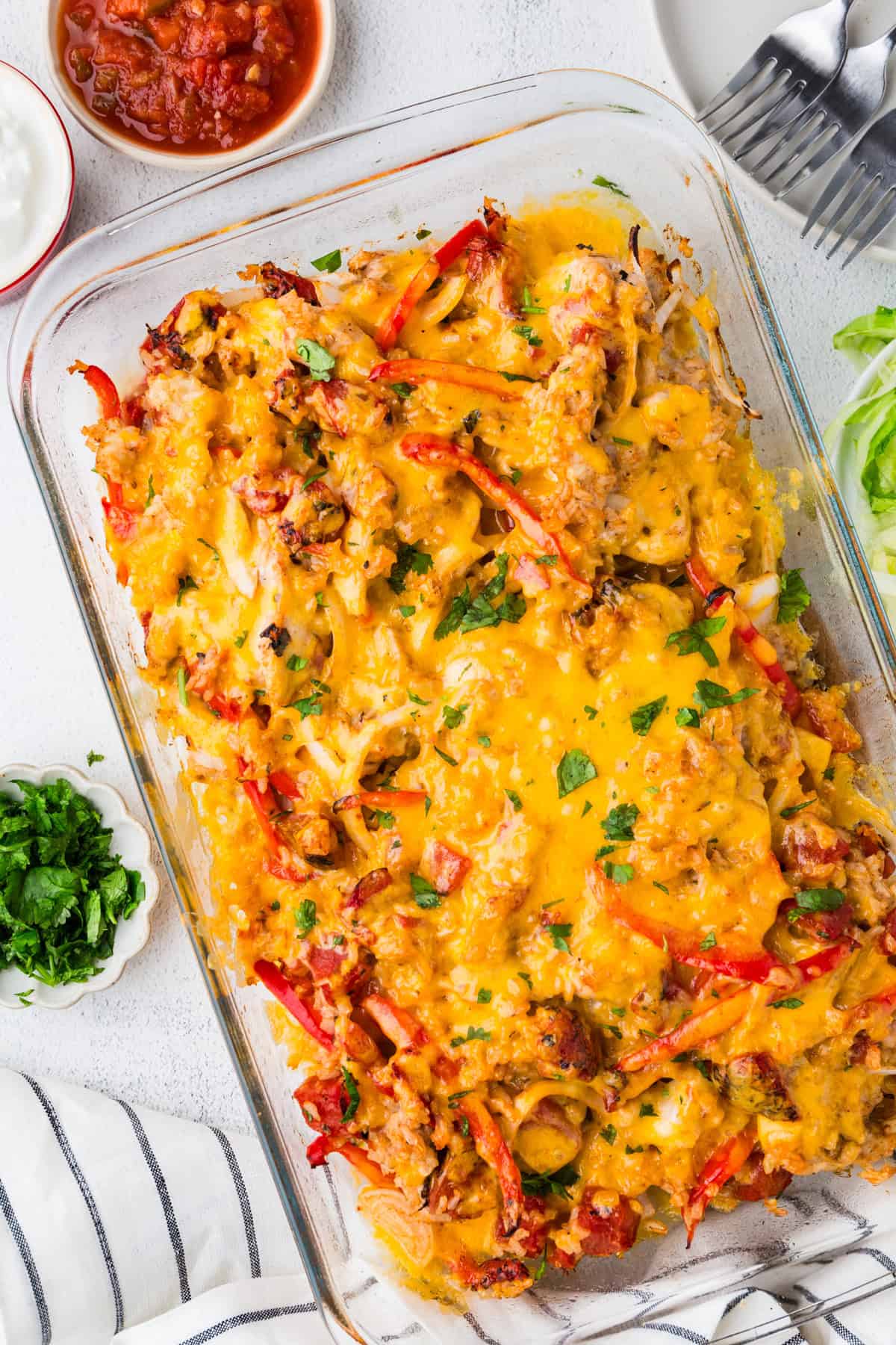 A baked cheesy chicken fajita casserole in a glass baking dish with bowls of salsa, sour cream and cilantro nearby as toppings.