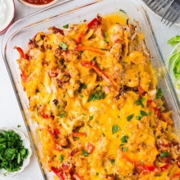 Close up of a baked cheesy chicken fajita casserole in a glass baking dish with bowls of salsa, sour cream and cilantro nearby as toppings.
