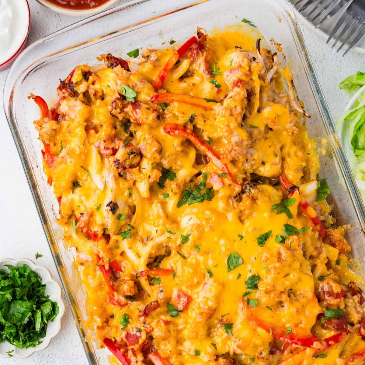 Close up of a baked cheesy chicken fajita casserole in a glass baking dish with bowls of salsa, sour cream and cilantro nearby as toppings.