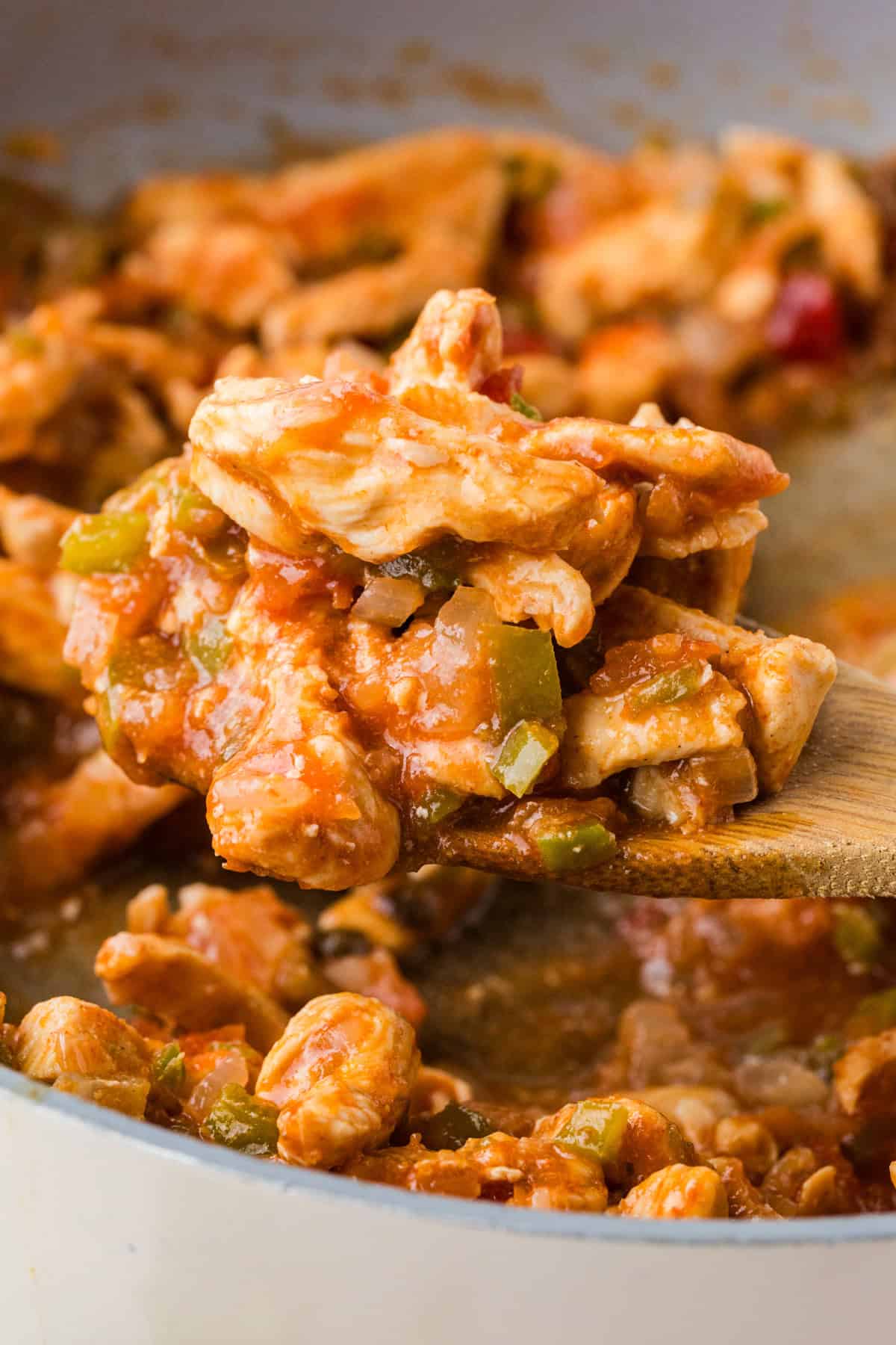 A wooden spoon lifting up a scoop of strips of chicken with onions, peppers and salsa from a skillet.