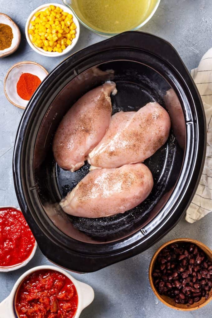 Seasoned chicken breasts in a slow cooker insert with bowls of other ingredients on the countertop nearby.