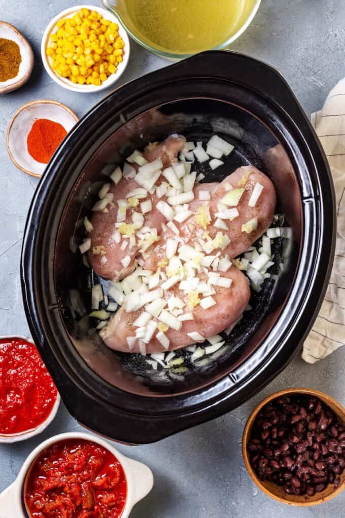 Seasoned chicken breasts in a slow cooker insert with chopped onion on top and bowls of other ingredients on the countertop nearby.