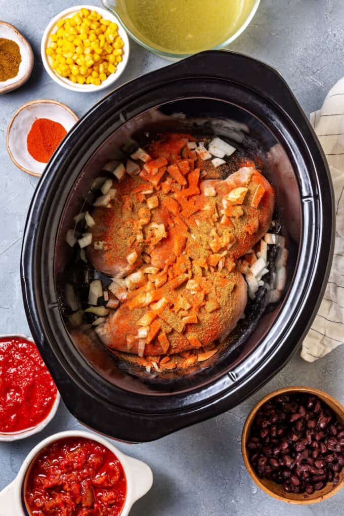 Seasoned chicken breasts in a slow cooker insert with chopped onion and seasonings on top and bowls of other ingredients on the countertop nearby.