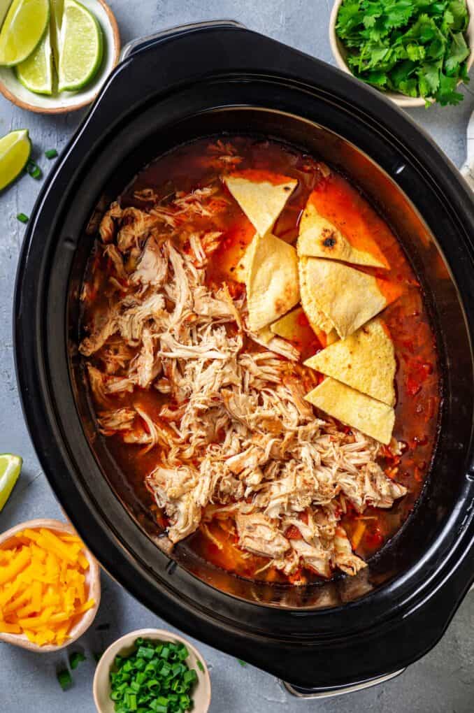 A slow cooker with shredded chicken and tortilla wedges being added into a soup.