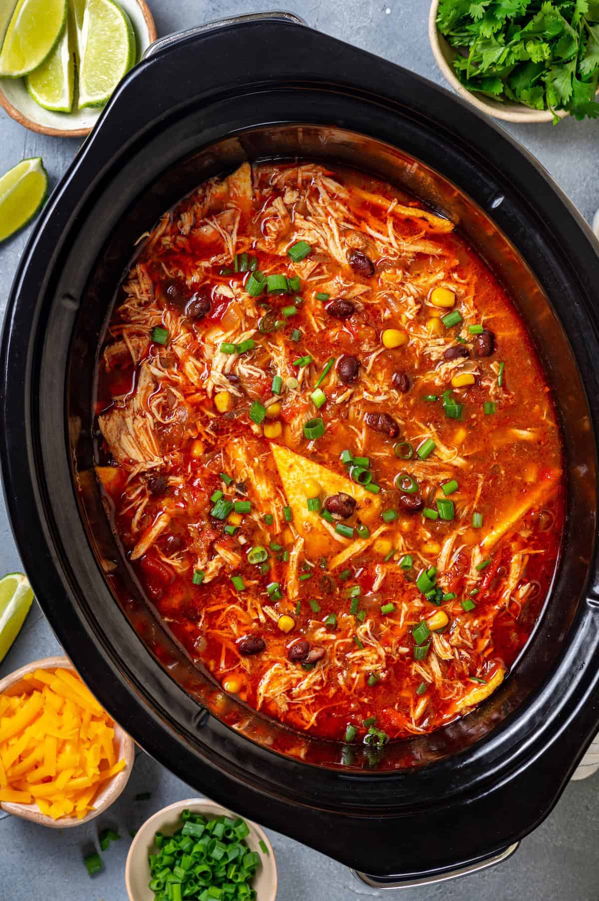 A slow cooker chicken enchilada soup with black beans, corn and tortillas and sliced green onions on top.