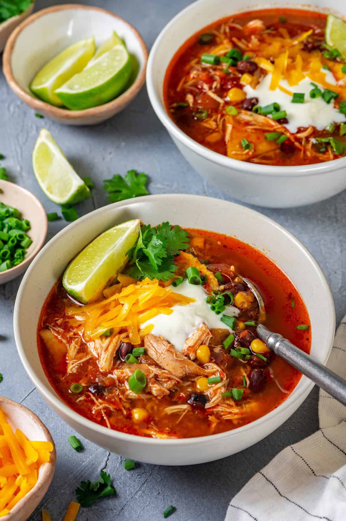 Bowls of chicken enchilada soup with spoon resting in them and sliced green onions, sour cream and shredded cheese on top.