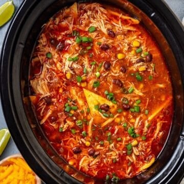 Close up of a slow cooker chicken enchilada soup with black beans, corn and tortillas and sliced green onions on top.