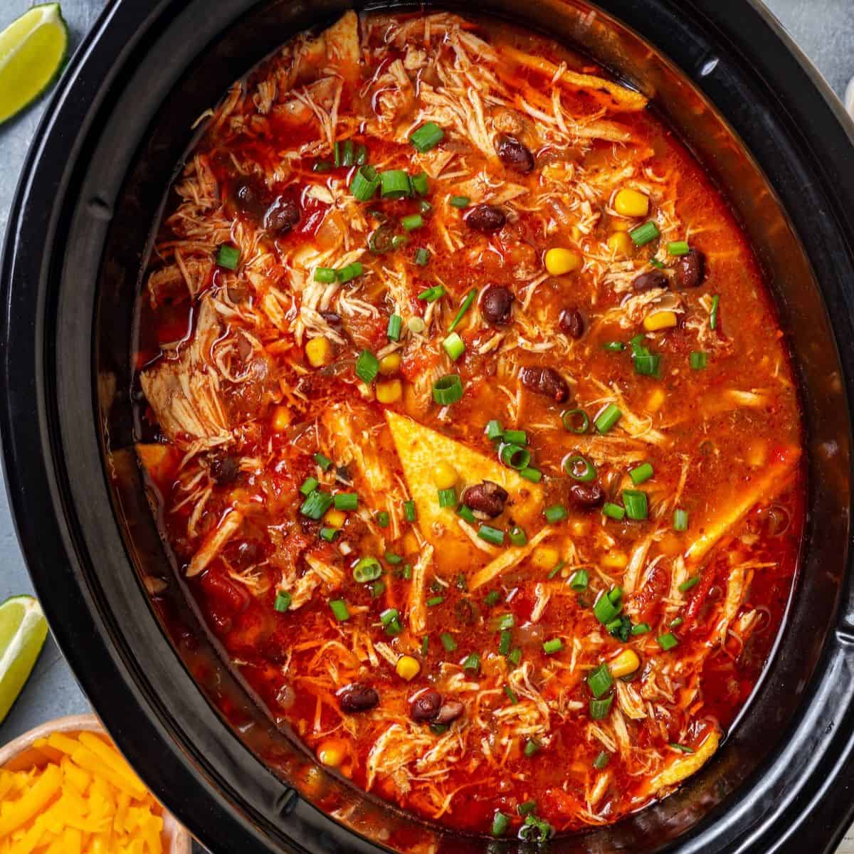 Close up of a slow cooker chicken enchilada soup with black beans, corn and tortillas and sliced green onions on top.