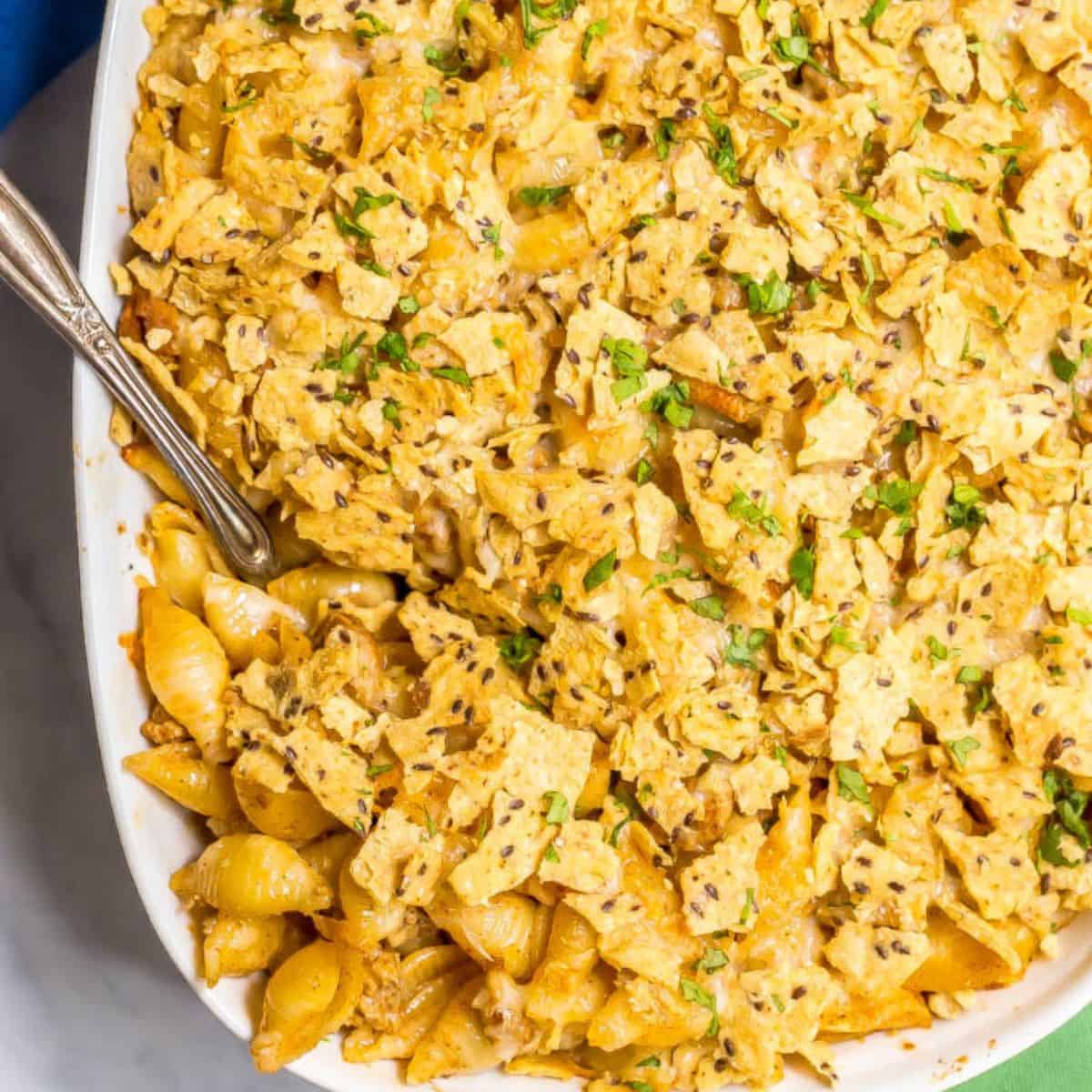 Close up of a serving spoon resting in a casserole dish with a taco mac and cheese topped with cheese and tortilla chips.