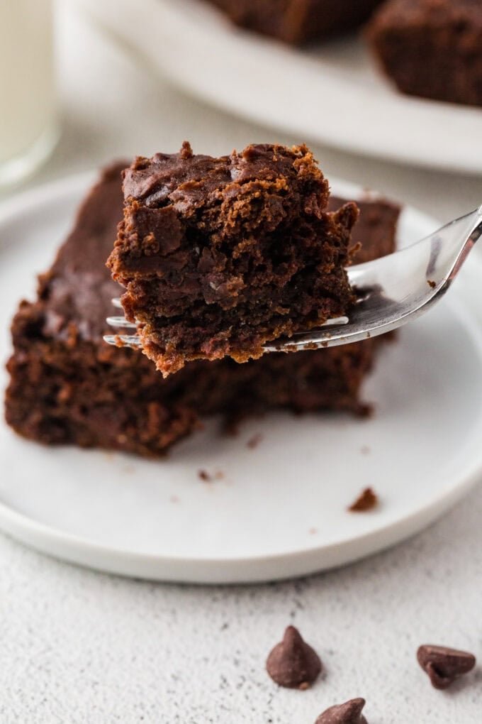 A fork holding up a bite of chocolate cake from a plate with a piece on it.