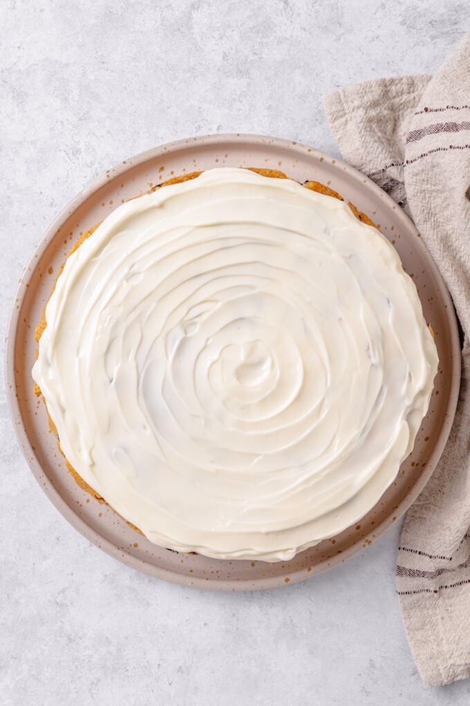 A round cake on a serving platter with cream cheese frosting spread on top.