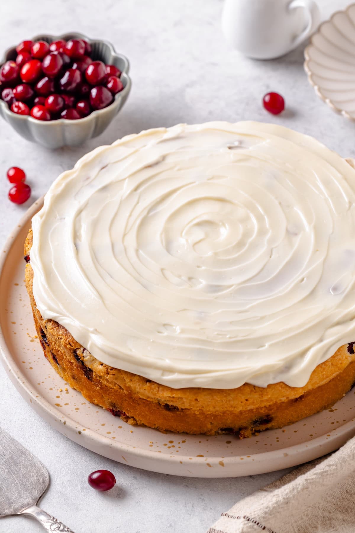 A fresh cranberry cake with cream cheese frosting on a serving plate with a bowl of fresh cranberries in the background.