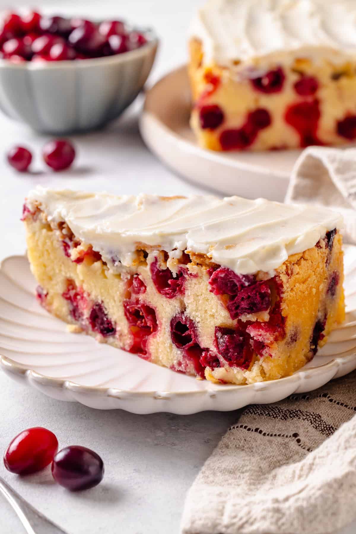 A slice of cranberry cake with cream cheese frosting on a small fluted round plate with cranberries scattered about.