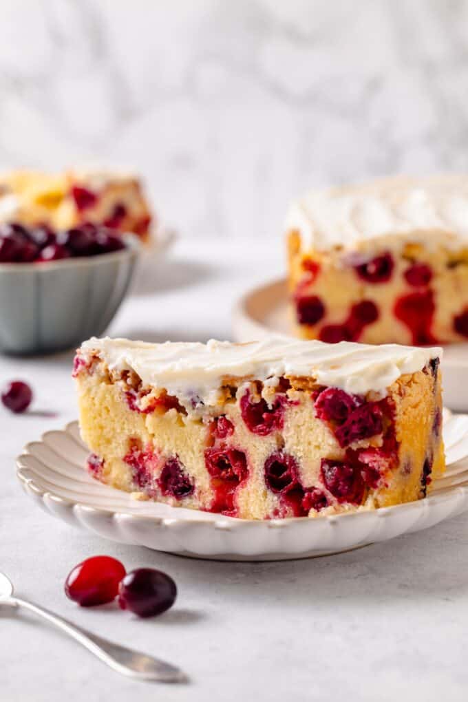 A slice of cranberry cake with cream cheese frosting on a small fluted round plate with the full cake in the background.