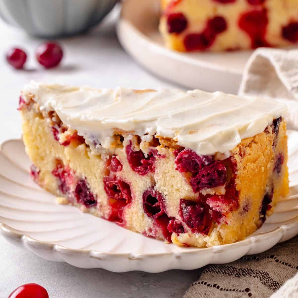Close up of a slice of cranberry cake with cream cheese frosting on a small fluted round plate with another slice in the background.