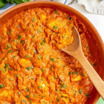 Close up of a wooden spoon resting in a copper skillet filled with a saucy chicken tikka masala mixture sprinkled with fresh parsley.