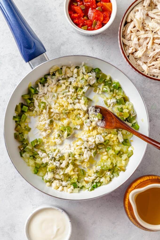 A wooden spoon stirring flour into a skillet with sautéed onion and green peppers.