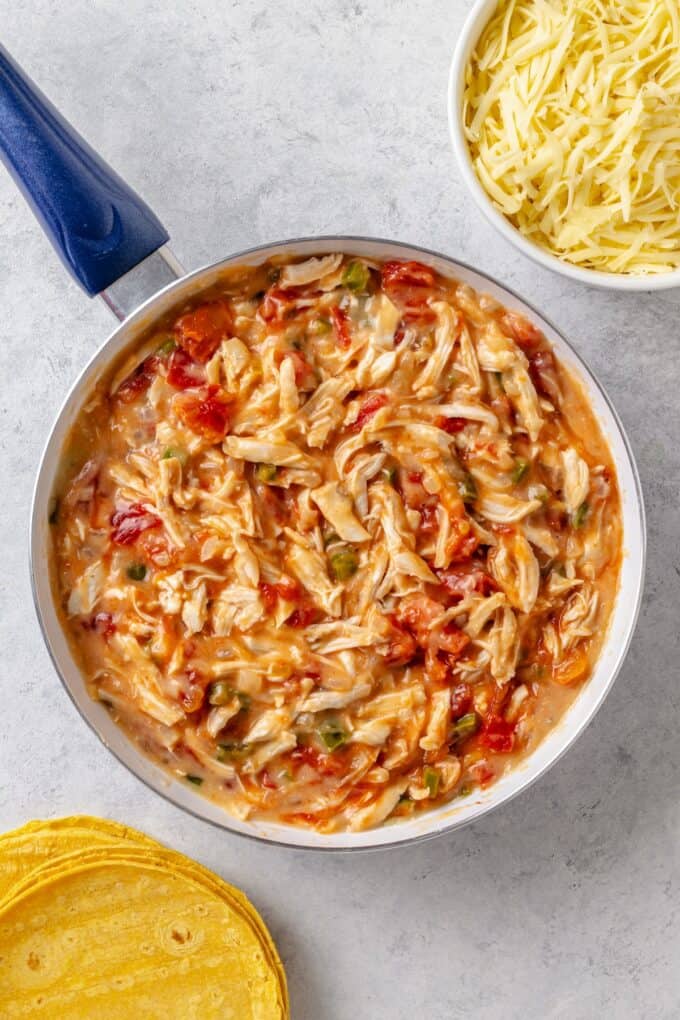 Shredded chicken and a can of Rotel stirred into a brothy mixture in a skillet with corn tortillas and a bowl of shredded cheese nearby.