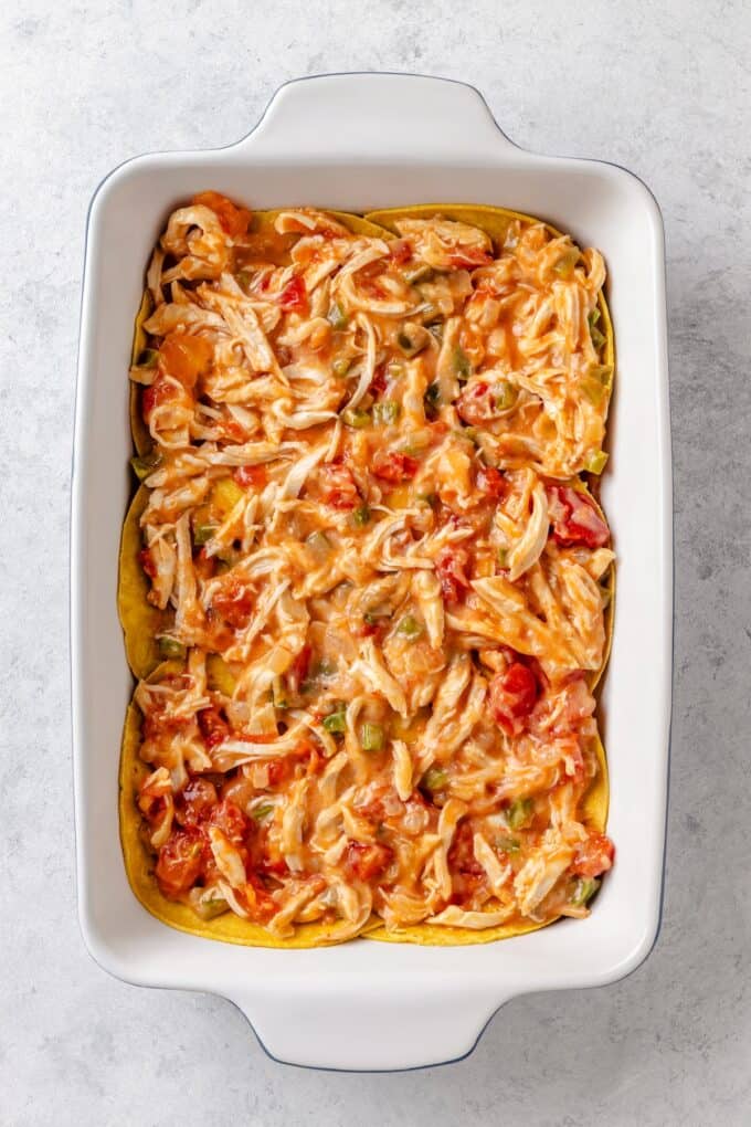 A creamy chicken and Rotel mixture with peppers and onions layered on top of corn tortillas in a casserole dish.