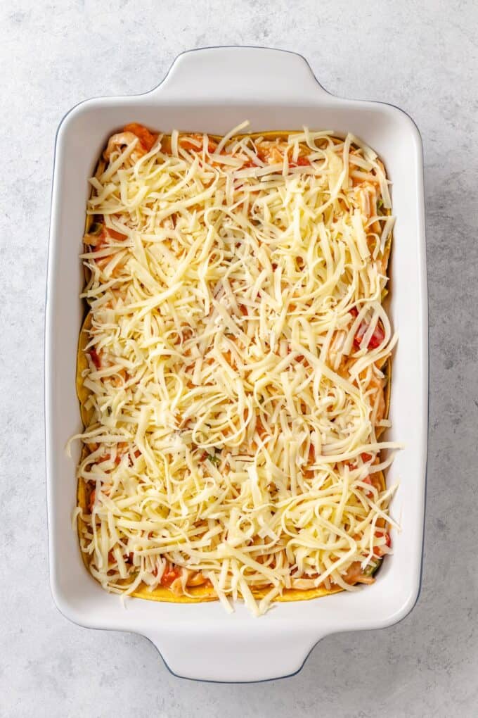 Shredded cheese sprinkled all over a creamy chicken and Rotel mixture with peppers and onions layered on top of corn tortillas in a casserole dish.