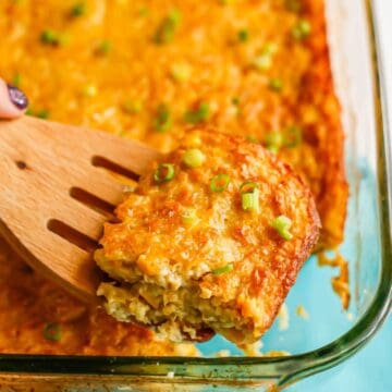 Close up of a wooden spatula lifting up a scoop of corn pudding from a clear baking dish.