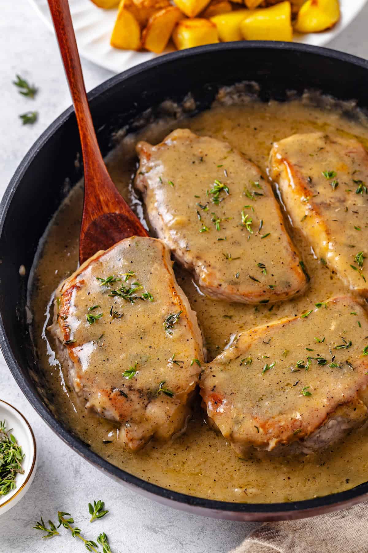 A spoon lifting up a pork chop wiht gravy and thyme from a skillet.