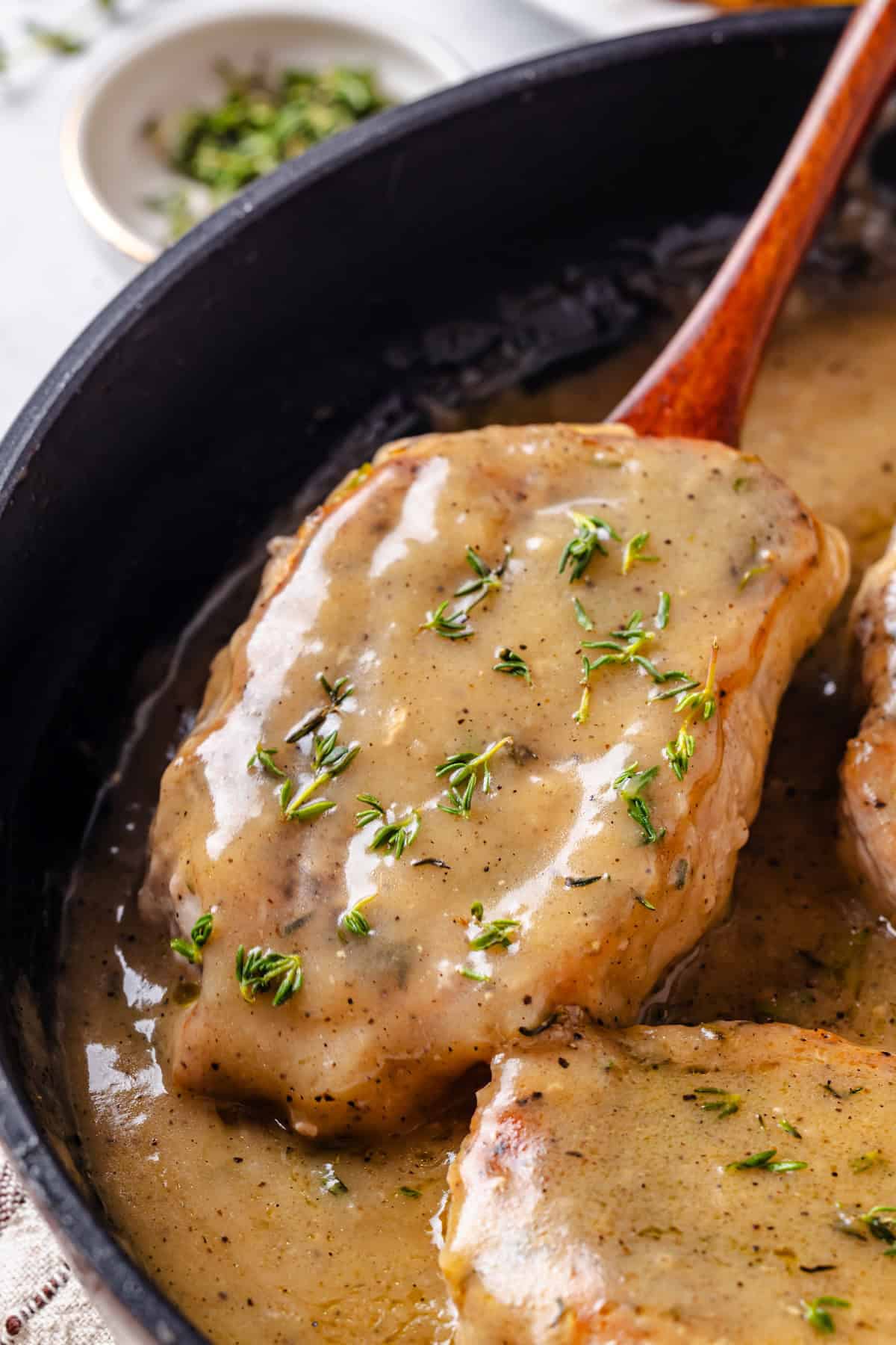 Close up of a wooden spoon lifting a pork chop with gravy and fresh thyme from a skillet.