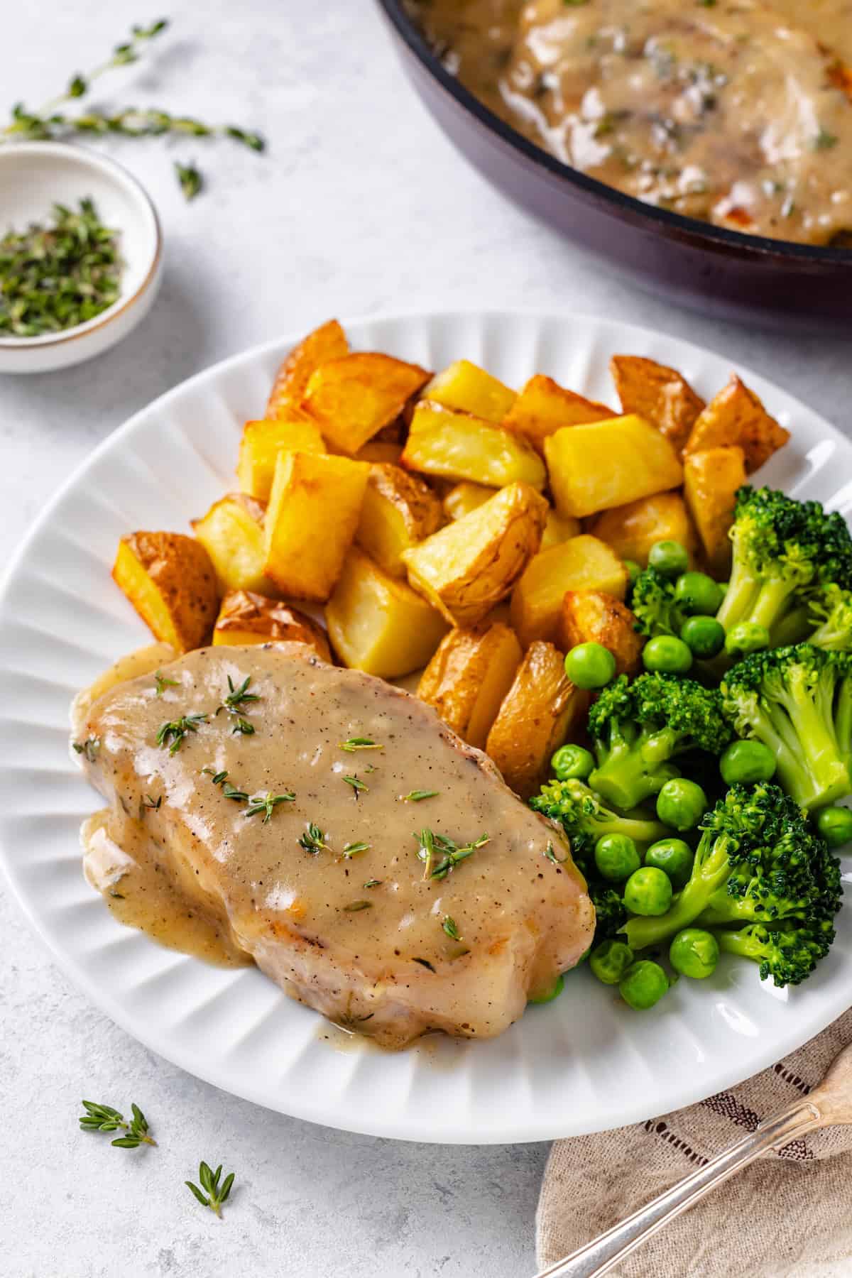 A dinner plate with a pork chop covered in gravy alongside roasted potatoes and a mixture of broccoli and green peas.