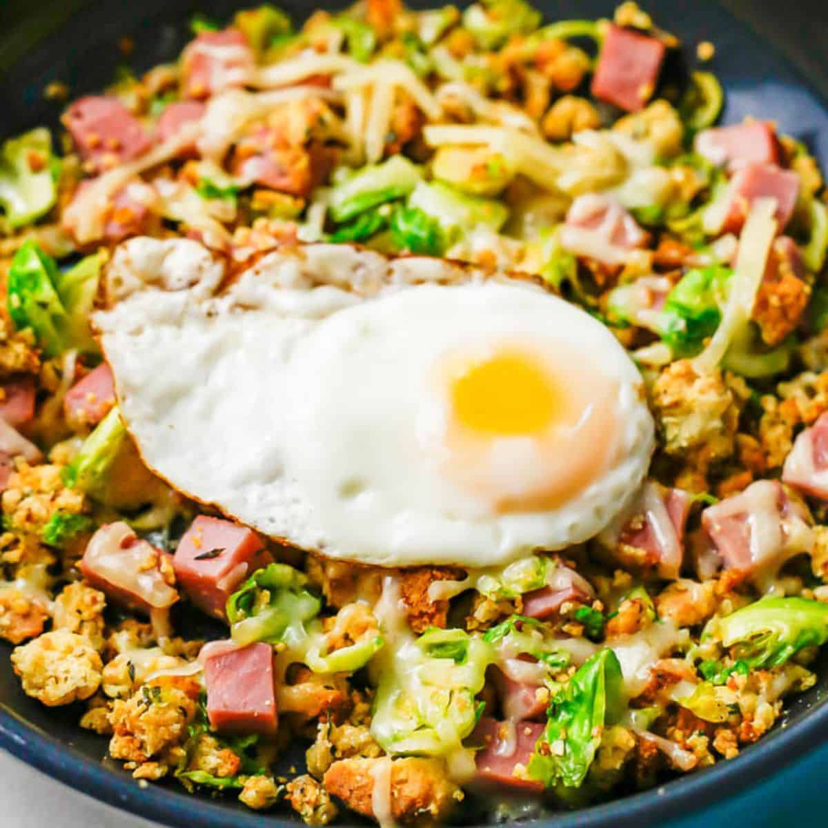 Close up of a fried egg on top of a breakfast hash with ham, stuffing and Brussels sprouts.
