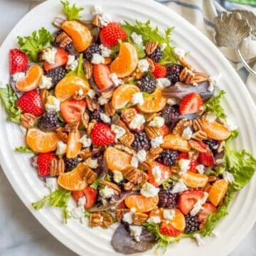 A large serving platter with a mixed greens salad topped with fresh fruit, nuts and creamy goat cheese.