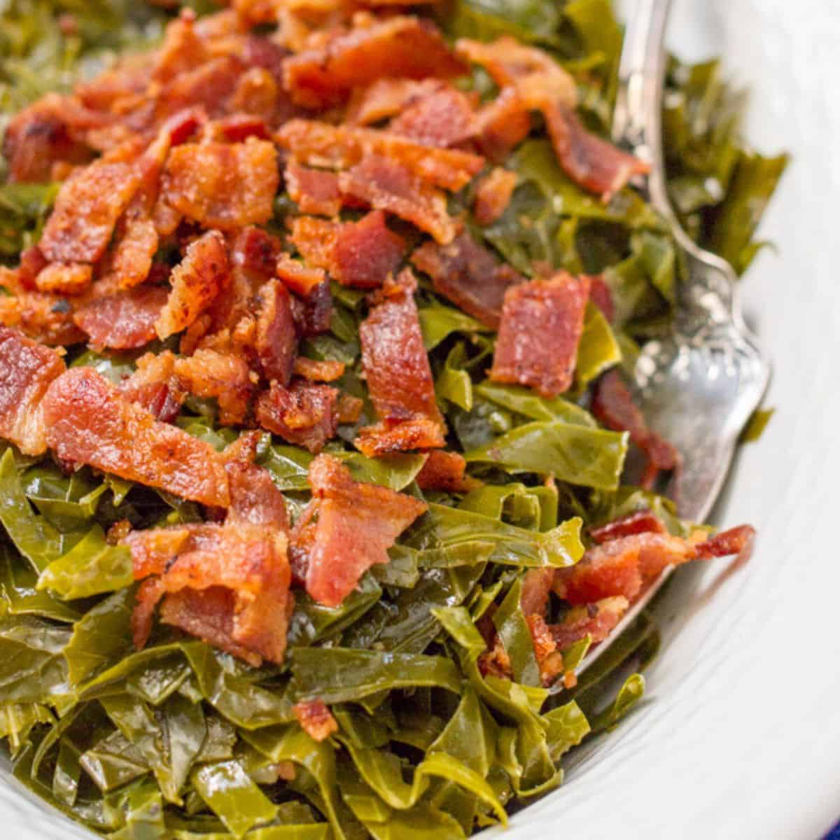 Close up of a serving bowl with sliced cooked collard greens topped with crisp, cooked bacon with a spoon resting in it.
