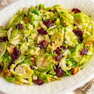 Close up of a white serving bowl full of shredded Brussels sprouts mixed with dried cranberries and chopped walnuts.
