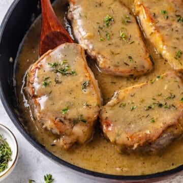 Close up of a spoon lifting up a pork chop wiht gravy and thyme from a skillet.