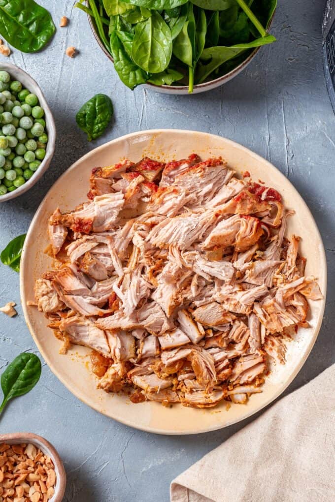 Shredded chicken on a plate with bowls of spinach and peas nearby.