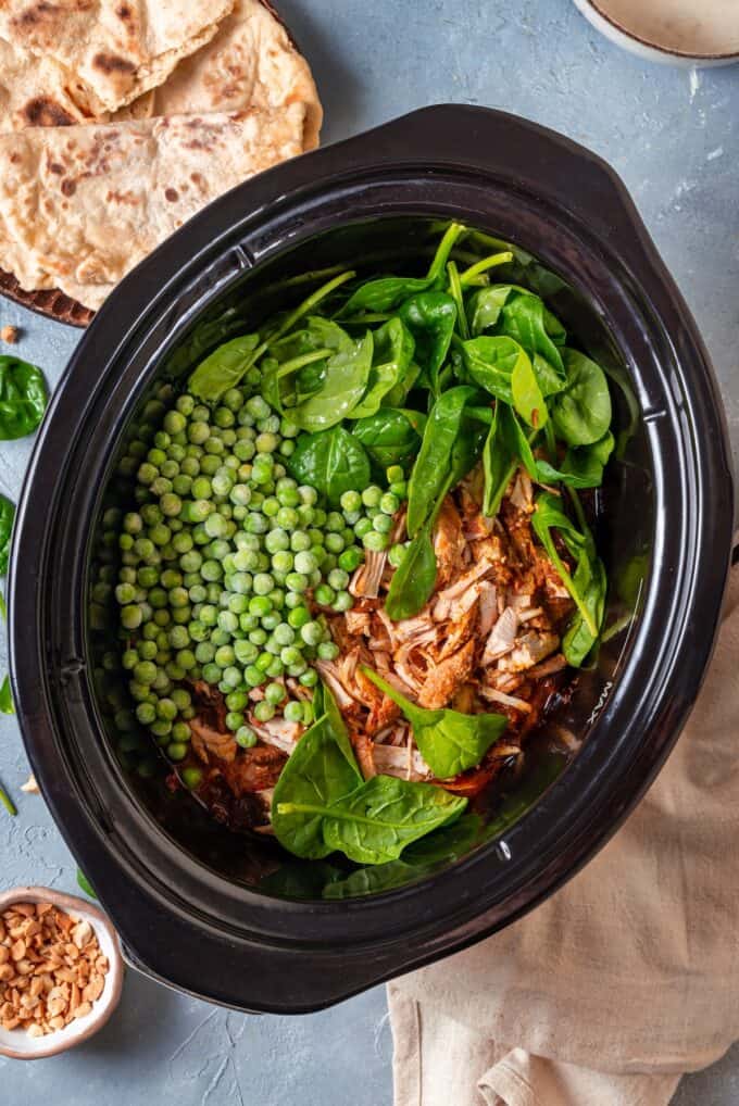 Frozen peas, fresh baby spinach and shredded chicken being added to a slow cooker before being stirred in.