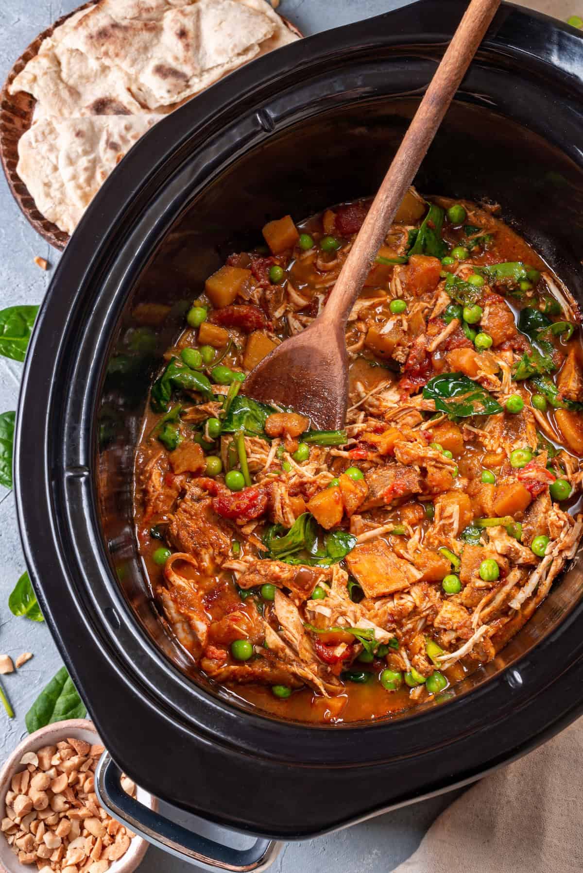 A wooden spoon resting in a slow cooker full of a chicken curry and veggie mix.