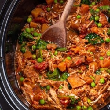 A slow cooker full of a chicken and sweet potato curry with a wooden spoon resting in it.