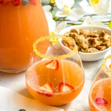 A stemless wine glass with a gold sugared rim and a champagne punch with fruit in it, with a pitcher in the background.