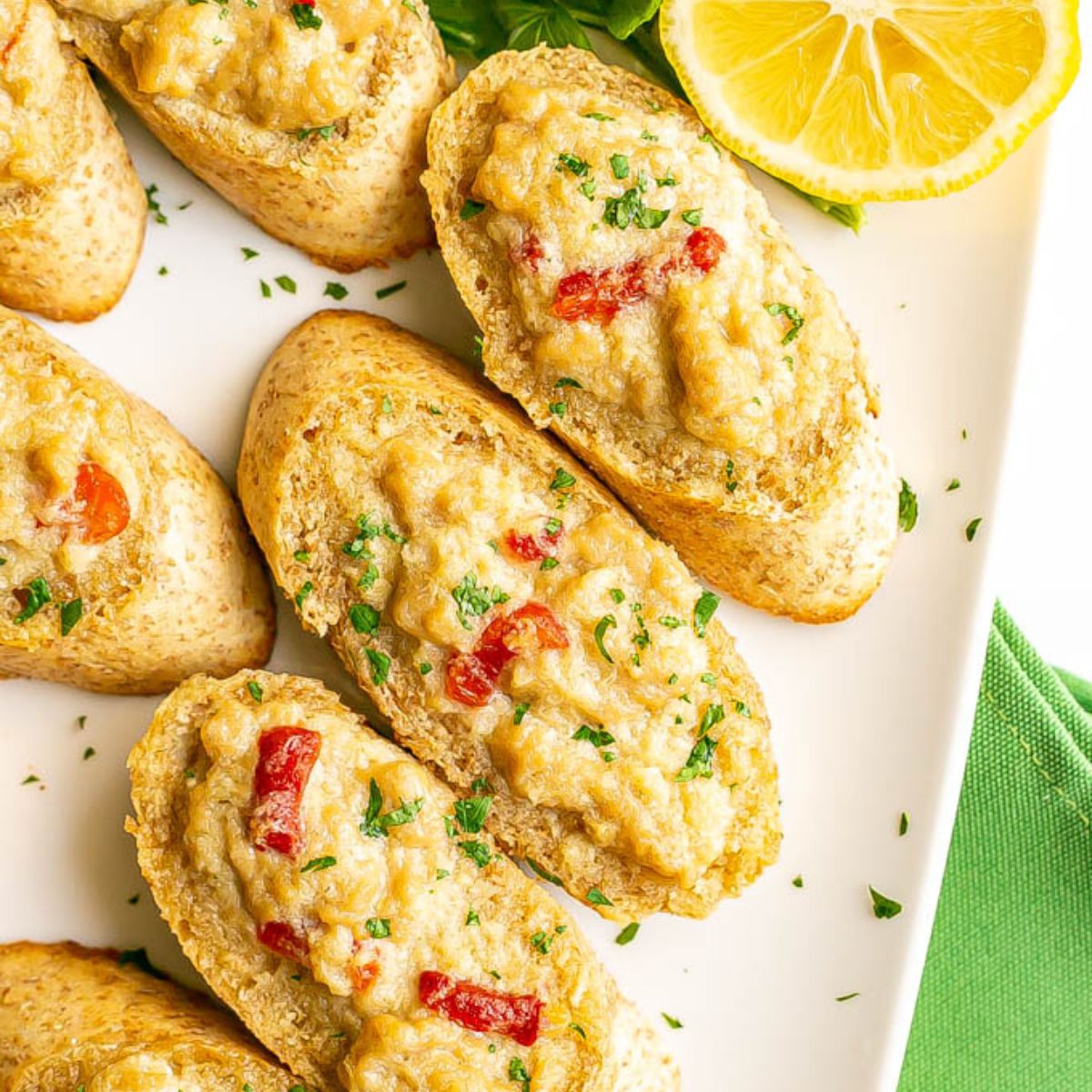 Close up of a white serving platter with crab melts on toasted baguette slices with a half lemon and fresh parsley to the side.