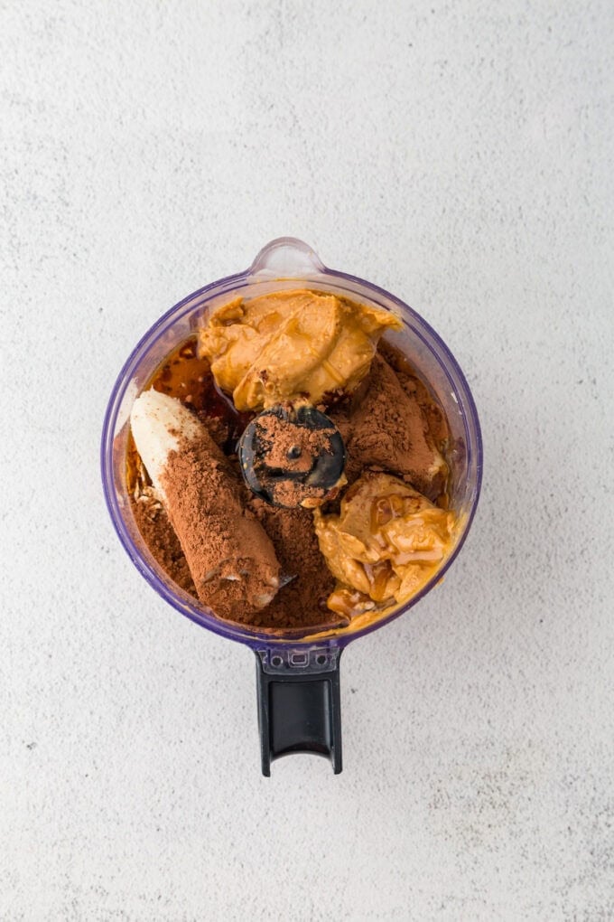 A food processor with ingredients layered in, including a banana, peanut butter and cocoa powder.
