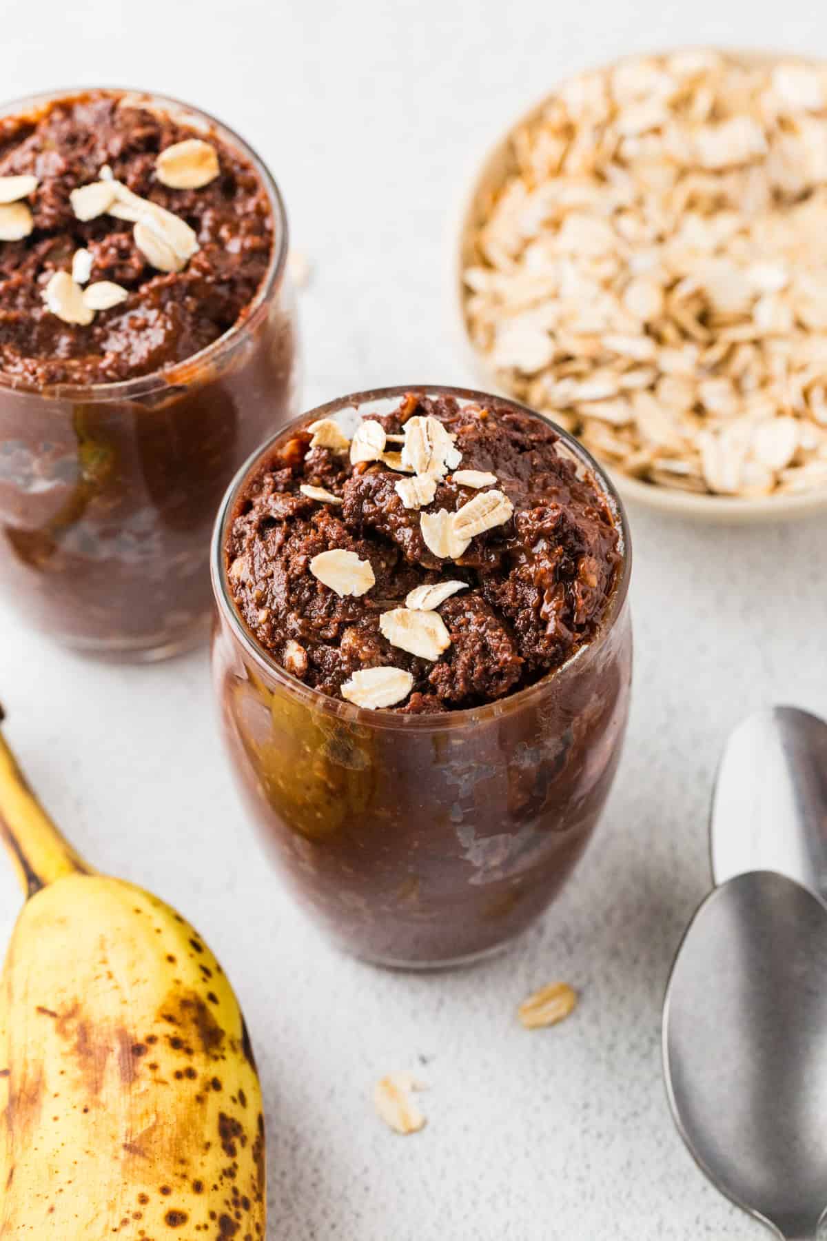 Top down look of a couple of glass jars filled with a fudgy chocolate pudding dessert with oats sprinkled on top and to the side, along with a ripe banana and two spoons.
