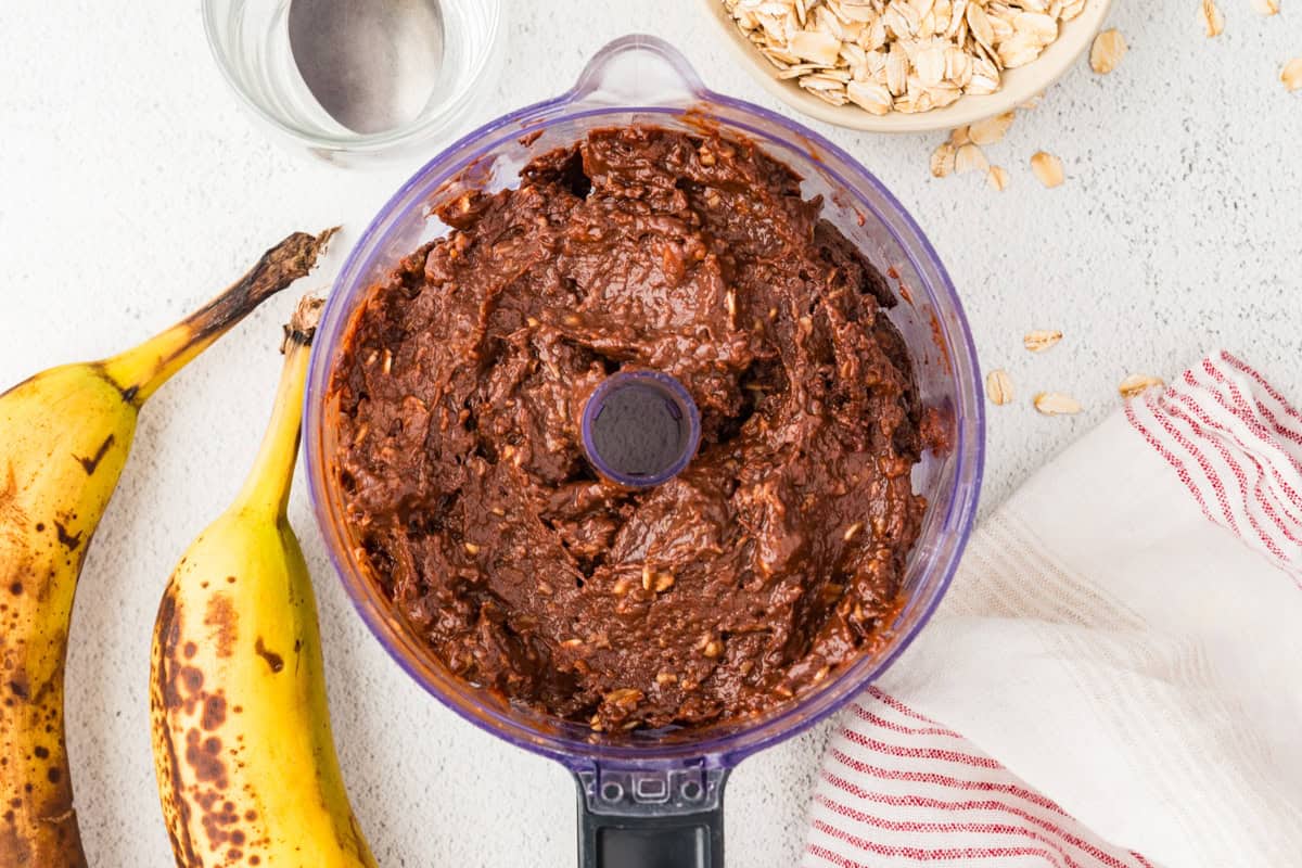 A chocolate pudding mixture in a food processor with a bowl of oats and a couple of ripe bananas to the side.