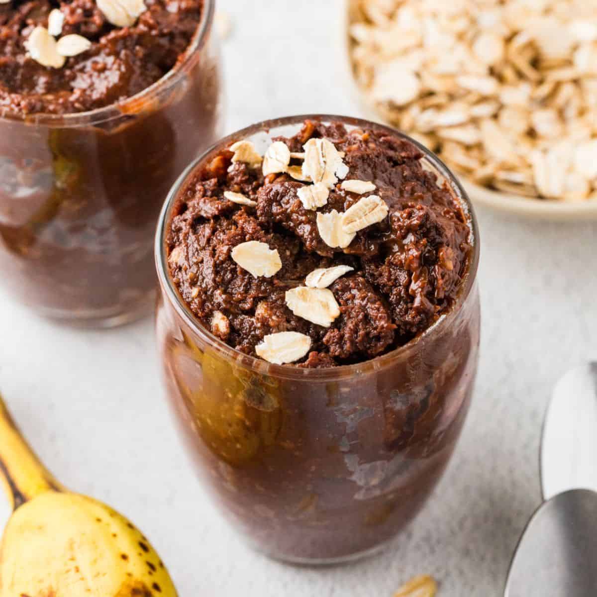 Close up of a couple of glass jars filled with a fudgy chocolate pudding dessert with oats sprinkled on top and to the side, along with a ripe banana and two spoons.