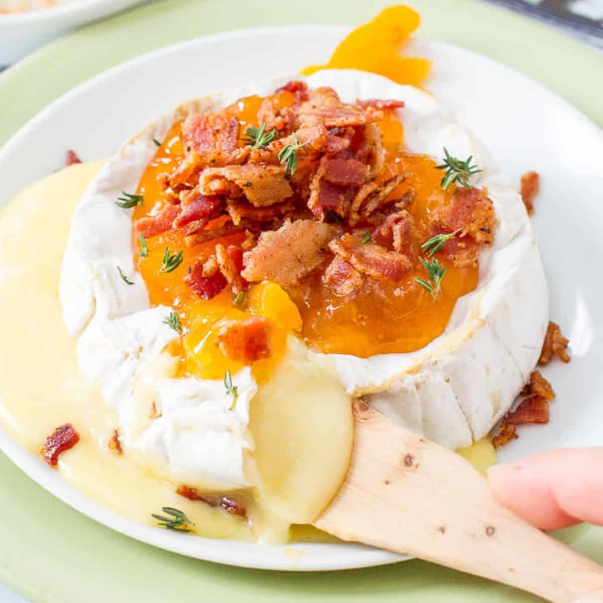 Close up of a small wooden cheese knife cutting into a wheel of baked brie cheese on a white plate with apricot jam and cooked, crumbled bacon on top.
