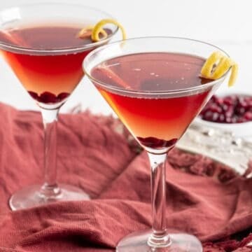 Close up of two pomegranate martinis with lemon twists on the side set on a burgundy kitchen towel.