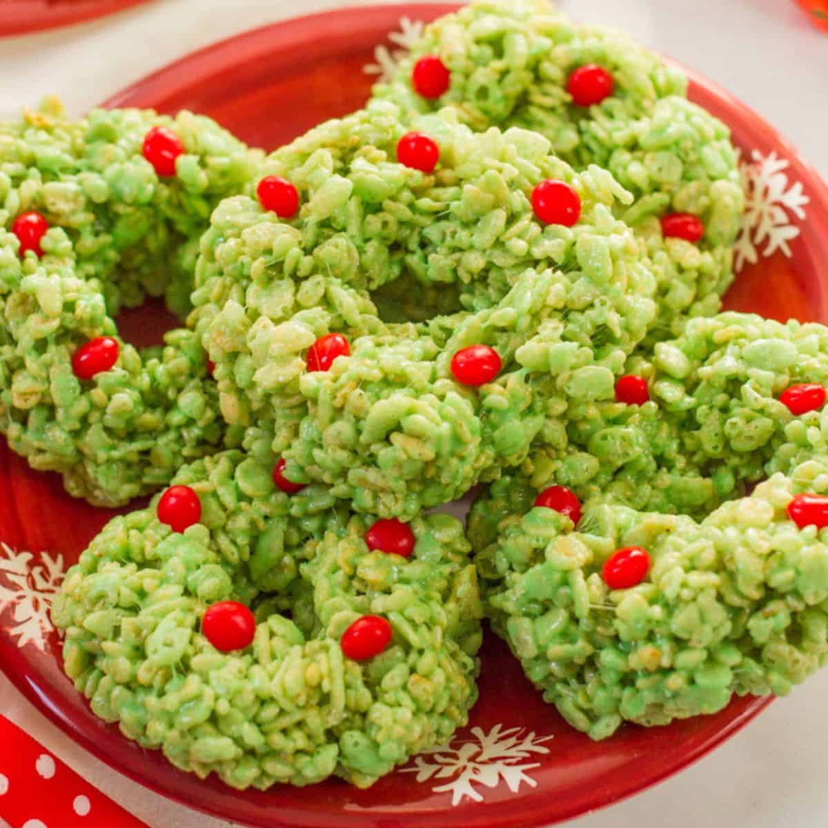 Close up of a red holiday plate with green Rice Krispie Christmas wreaths with small red hot candies on top arranged on it.