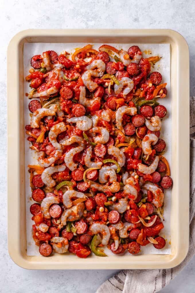 A parchment paper lined sheet pan with a mix of sausage and veggies with shrimp being added but not yet coooked.