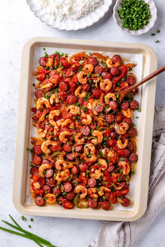 A wooden spoon resting on a sheet pan filled with a shrimp and sausage jambalaya mix.