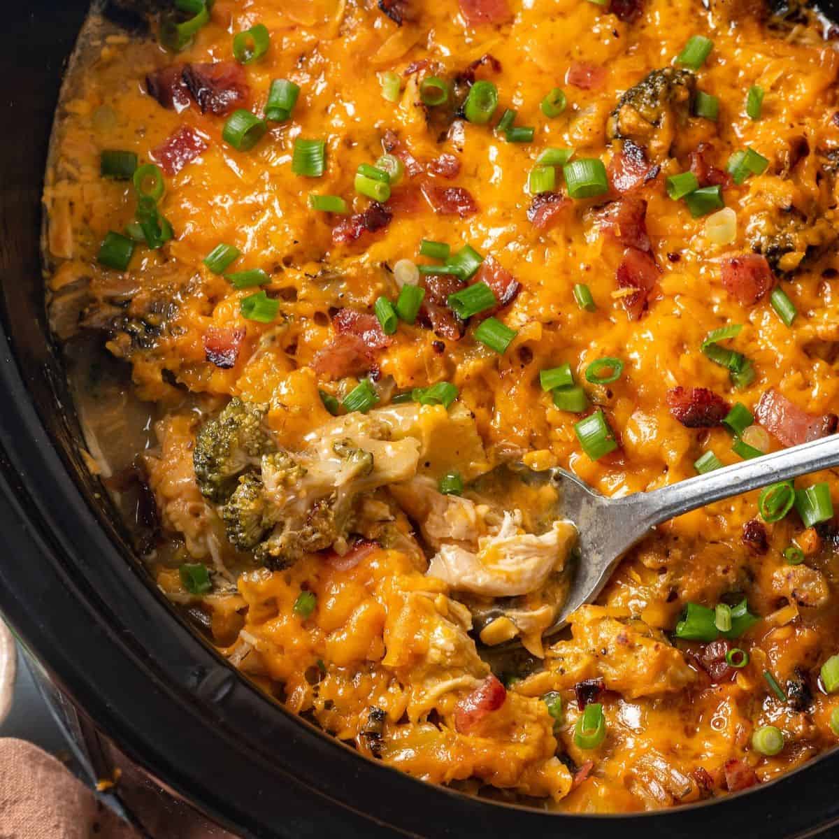 Close up of a serving spoon scooping up some cheesy chicken casserole with potatoes and broccoli from a slow cooker insert.